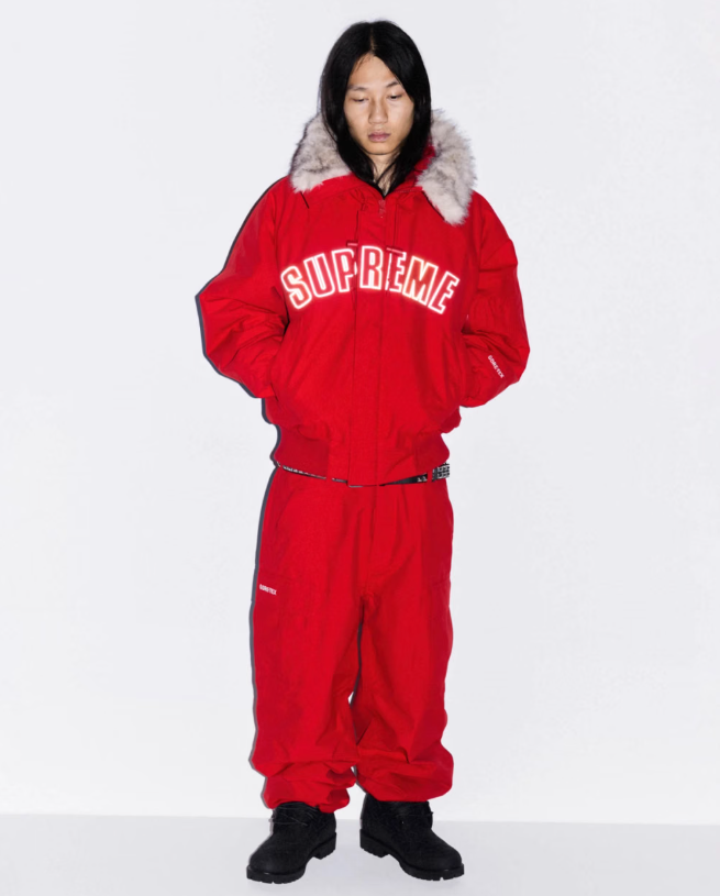 A person wearing a red Supreme hooded jacket with fur trim, standing against a white background.