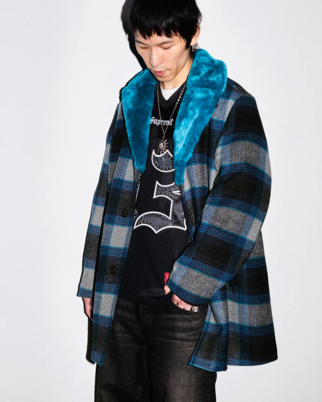 A person wearing a blue fur-trimmed plaid coat, a black t-shirt with a graphic design, and black pants.
