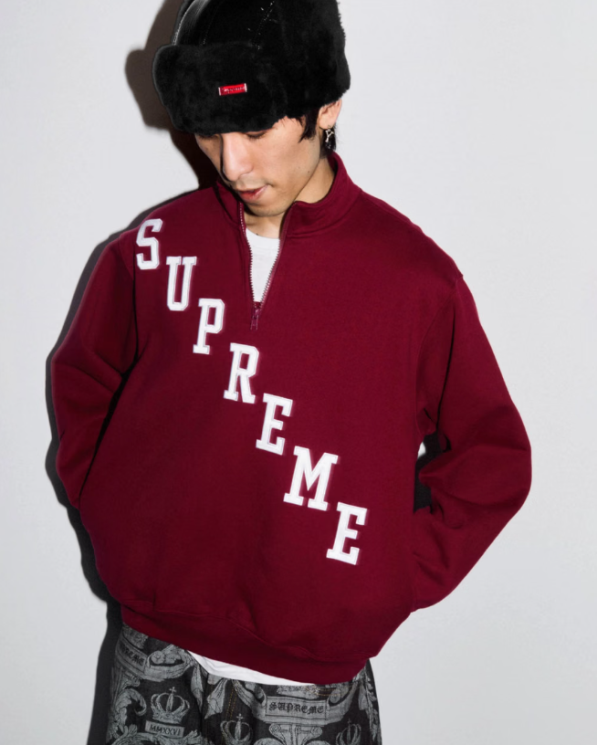 A person wearing a red Supreme hoodie and a black furry hat, standing against a white background.