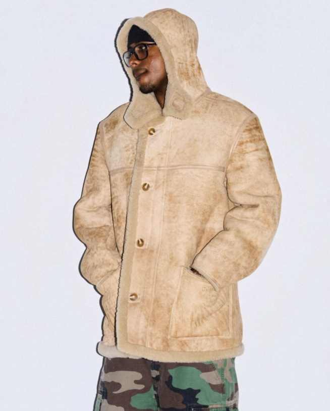 A person wearing a beige hooded jacket and sunglasses standing in front of a plain background.