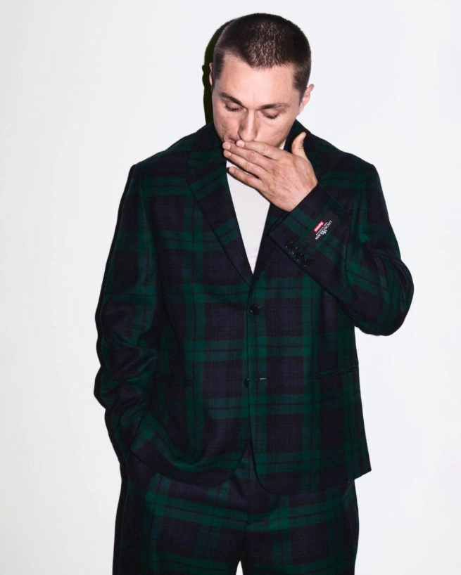 A man in a green and black plaid coat covering his mouth with his hand, looking down with a pensive expression.