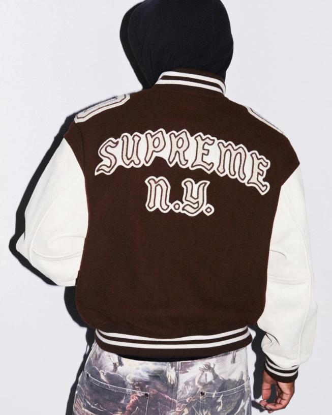 A person wearing a brown and white letterman-style jacket with the text "SUPREME N.Y." on the back.