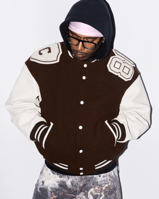 A person wearing a black hooded sweatshirt, glasses, and a brown and white varsity-style jacket.