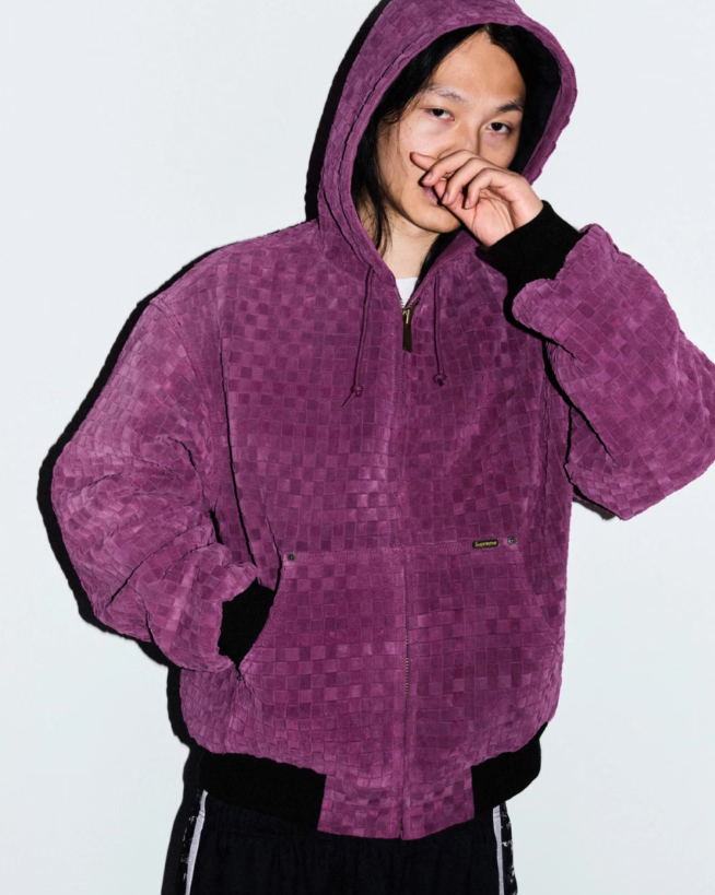 A person wearing a purple hooded jacket with a checkered pattern, standing against a white background and covering their face with their hand.