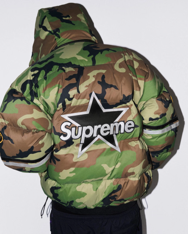 A person wearing a camouflage jacket with a large Supreme logo on the back.