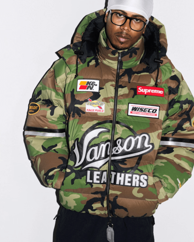 A person wearing a camouflage jacket with various brand logos and patches, including Supreme, K&N, Wiseco, and Van&son Leathers.