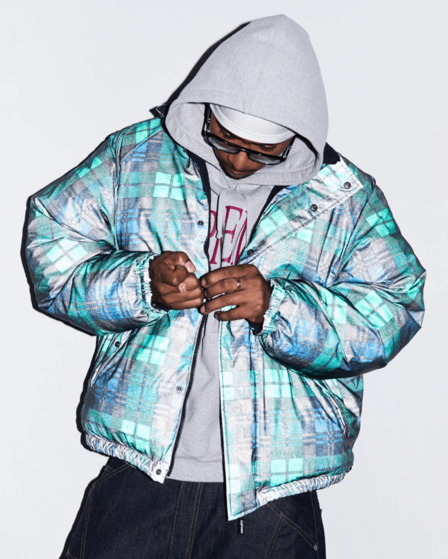 A person wearing a hooded jacket with a colorful, patterned design stands with their hands clasped in front of them.