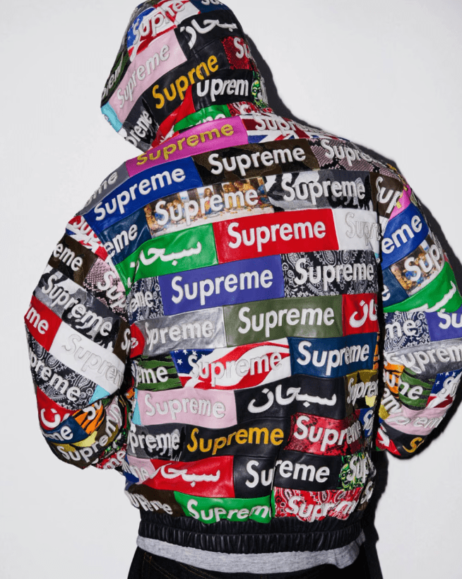 A person wearing a hooded jacket covered in the Supreme brand logo in various colors and sizes.