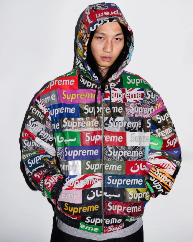 A person wearing a hooded jacket covered in the Supreme brand logo in various colors and languages.