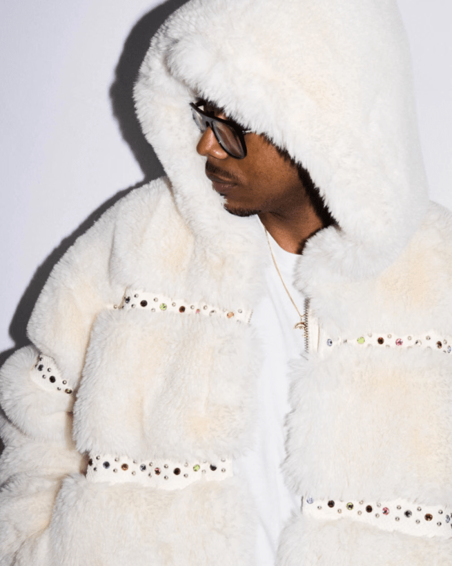 A person wearing a white hooded fur coat with studded details, looking down with a serious expression and wearing dark sunglasses.