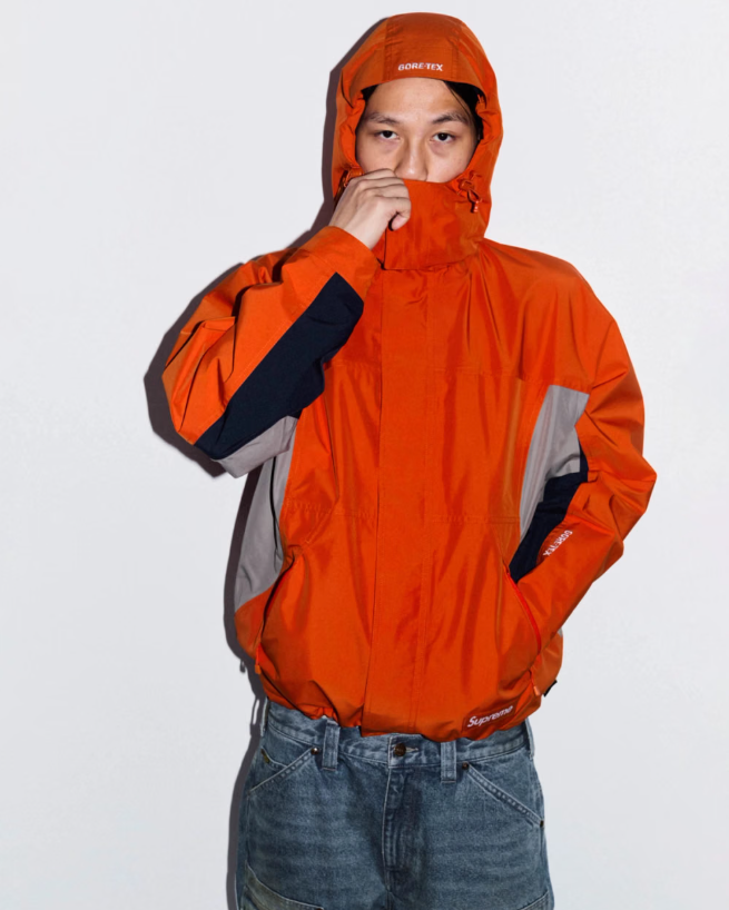 A person wearing an orange Gore-Tex jacket with a hood pulled up, partially covering their face.