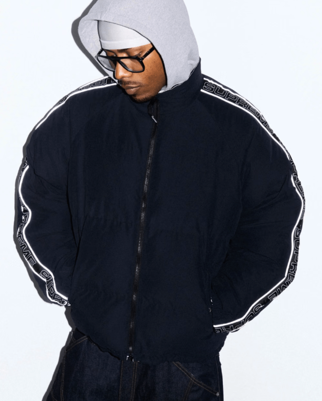 A person wearing a black jacket with white stripes and a gray hooded sweatshirt, standing against a white background and wearing sunglasses.