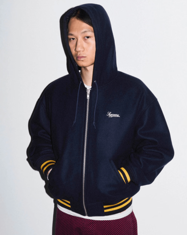 A person wearing a black hooded jacket with yellow stripes on the sleeves, standing against a plain white background.