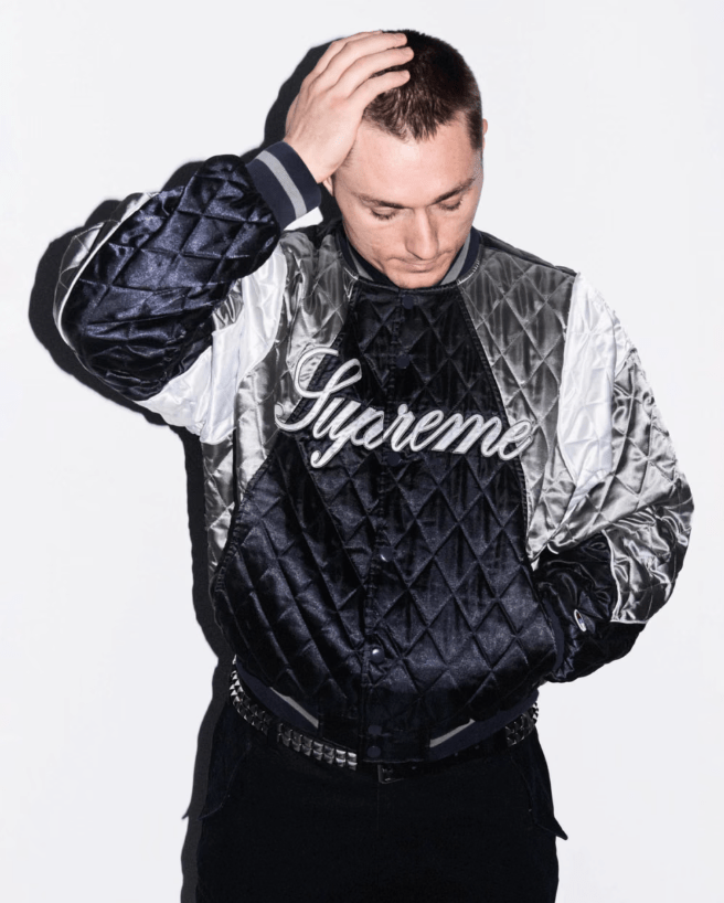 A person wearing a black and silver quilted jacket with the Supreme logo, holding their hand to their head in a pensive gesture against a white background.