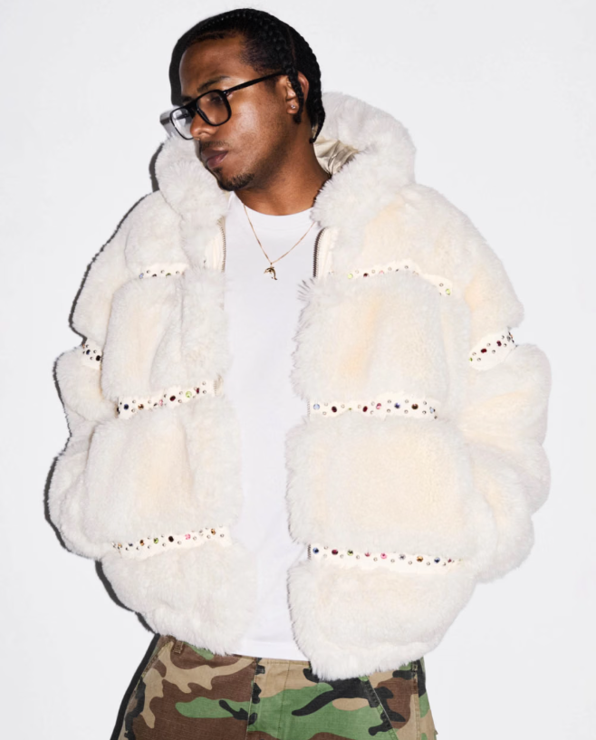 A person wearing a white furry coat with decorative patterns, standing against a white background and wearing glasses.