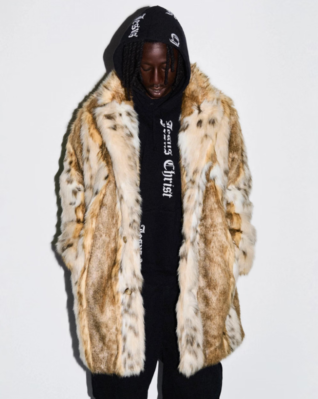 A person wearing a black hooded sweatshirt and a furry tan coat standing against a white background.
