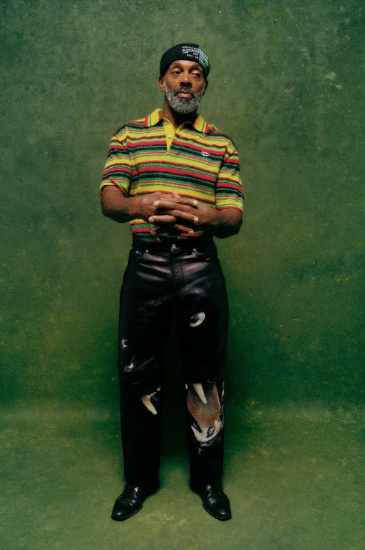 A man with a beard wearing a colorful striped shirt and a black hat, standing in front of a green background.