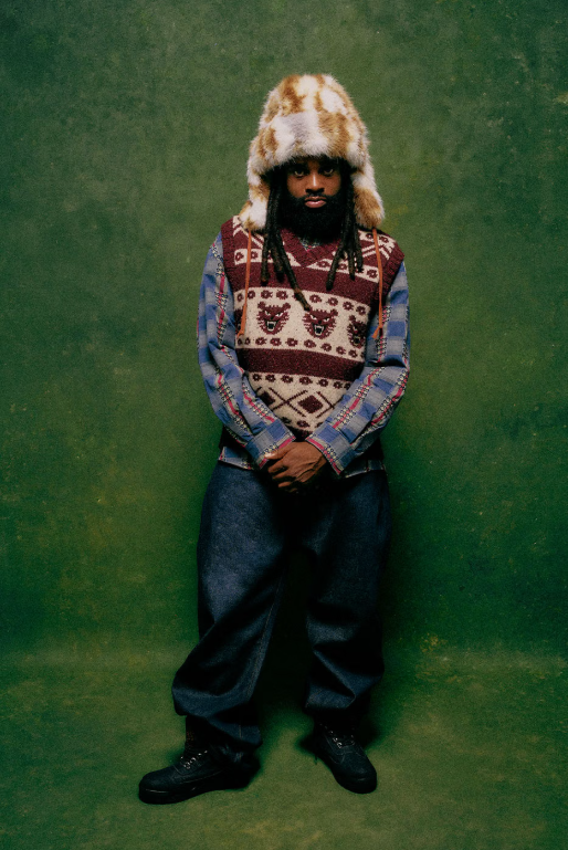 A person wearing a fur-trimmed hat and patterned sweater standing against a green background.