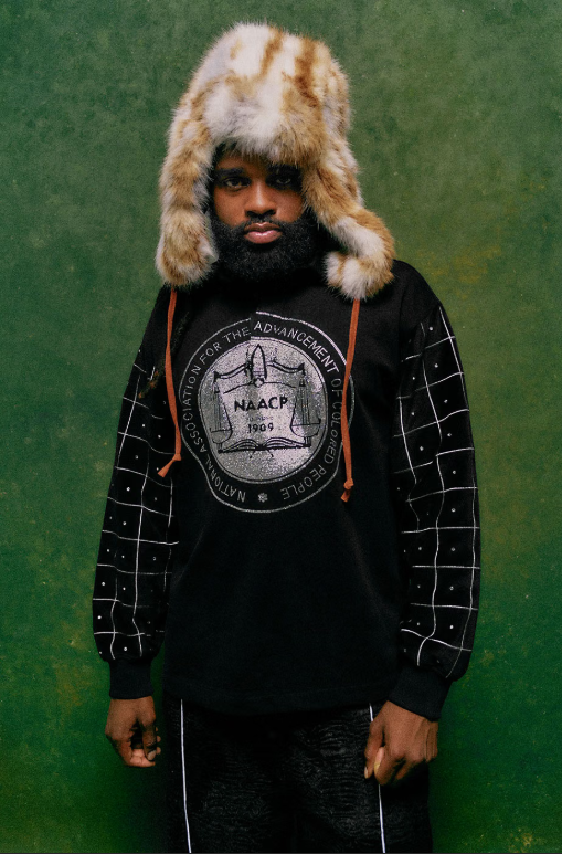 A person wearing a fur-lined hat and a black sweatshirt with a graphic design stands in front of a green background.