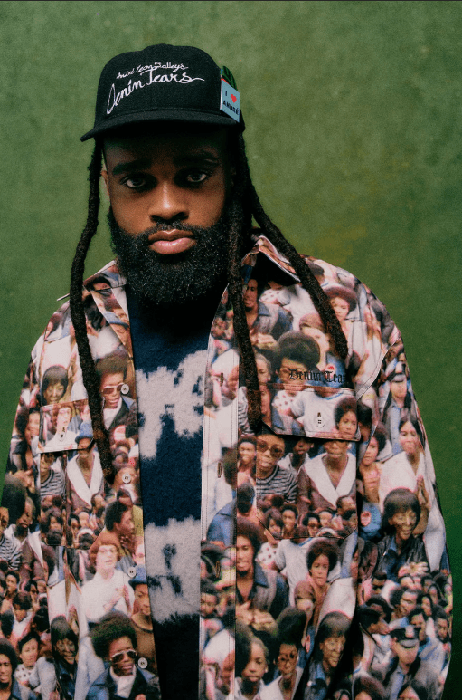 A person wearing a black hat with the text "Lemin Leaks" and a patterned shirt with many faces printed on it, standing against a green background.