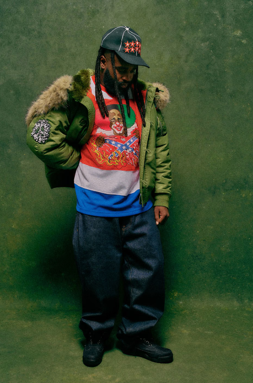 A person wearing a green jacket with fur trim, a baseball cap, and a colorful graphic t-shirt standing against a green background.