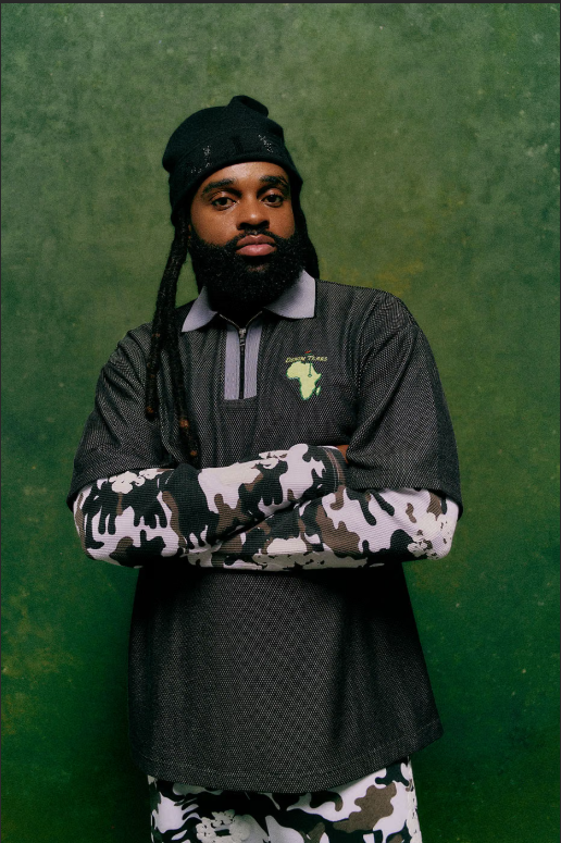 A man with dreadlocks and a beard wearing a black hat, camouflage jacket, and black shirt with an Africa logo, standing with his arms crossed against a green background.