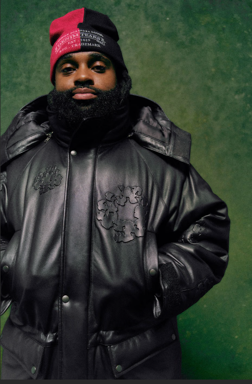 A man wearing a red and black hat and a black leather jacket stands against a green background.