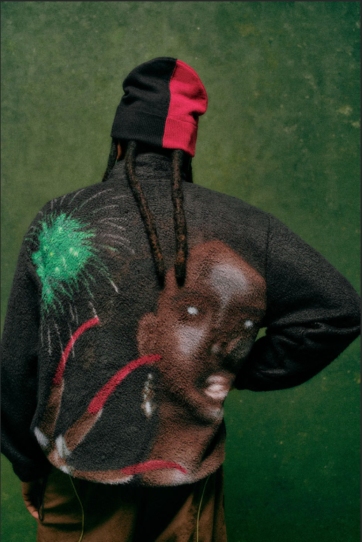 A person wearing a black and red hooded jacket with a colorful graphic design on the back, standing against a green background.