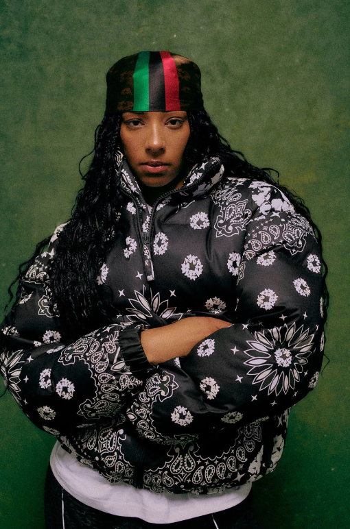 A person wearing a black and red patterned hoodie and a black and green headband, standing against a green background.