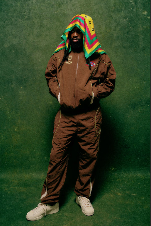 A person wearing a colorful hooded jacket and standing in front of a green background.