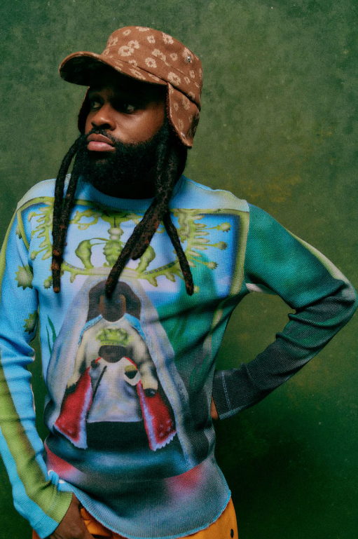 A person wearing a patterned hat and a colorful graphic sweatshirt, with dreadlocks, standing in front of a green background.