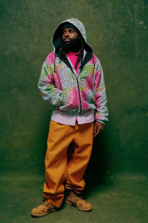 A person wearing a colorful hooded jacket and pants standing in front of a green background.