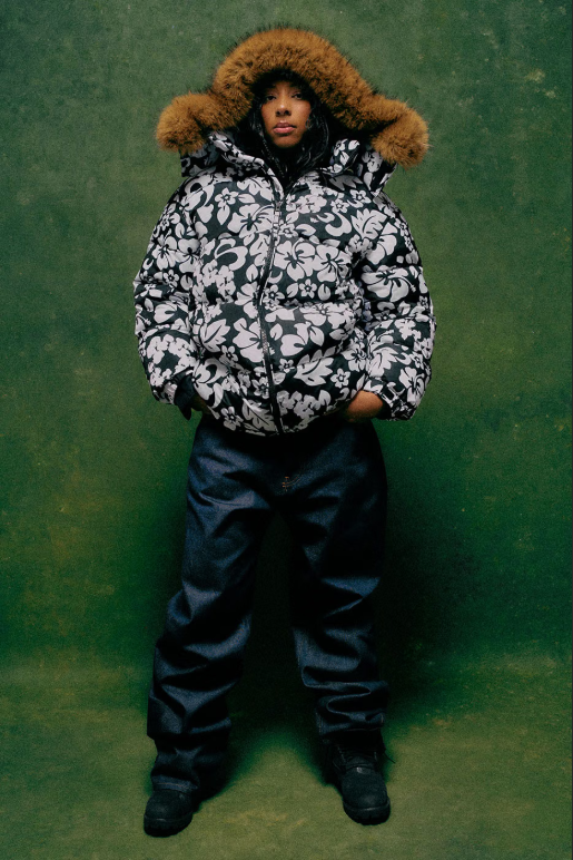 A person wearing a hooded, floral-patterned winter jacket with a fur-trimmed hood, standing against a green background.