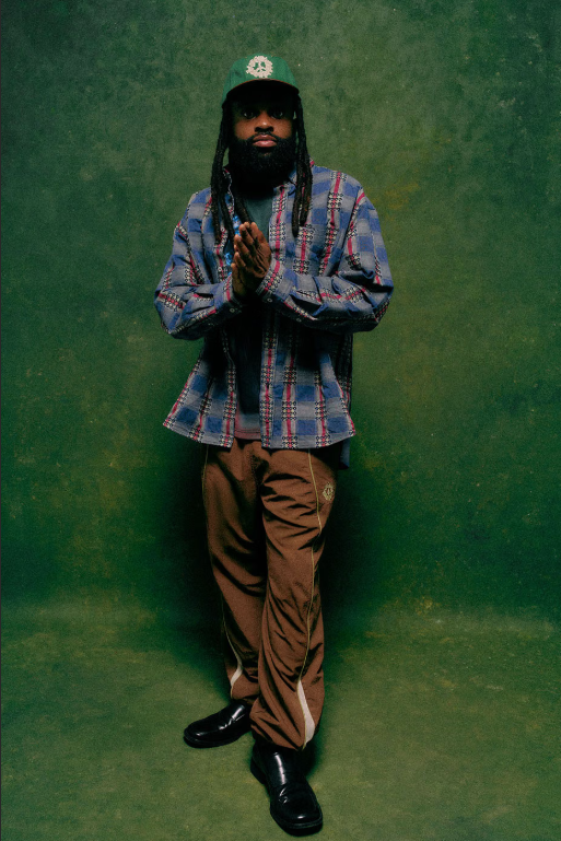 A person with a long beard and dreadlocks wearing a plaid shirt and brown pants, standing with their hands clasped in front of them against a green background.