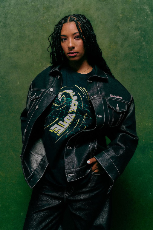 A young woman with braided hair wearing a black leather jacket and a graphic t-shirt stands against a green background.