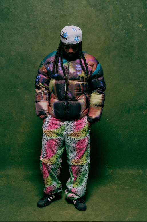 A person wearing a colorful, patterned jacket and hat standing in front of a green background.