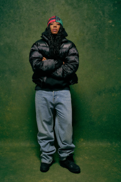 A person wearing a black puffy jacket and a red and blue hat standing against a green background.