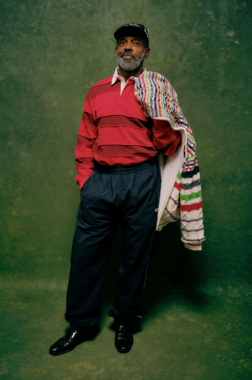 A man with a beard wearing a red and white striped shirt and holding a colorful patterned shawl, standing in front of a green background.