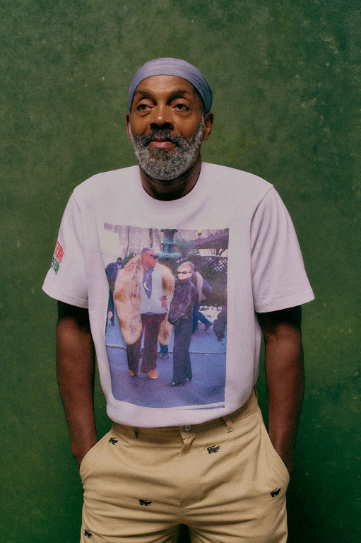 A bearded man wearing a white t-shirt with a printed image on it, standing in front of a green background.