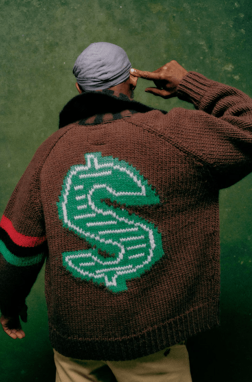 A person wearing a brown sweater with a green dollar sign logo, standing in a grassy area.