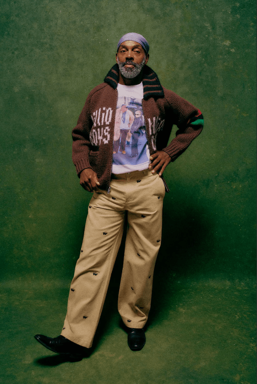 A man with a beard wearing a brown sweater with a graphic design and beige pants, standing against a green background.