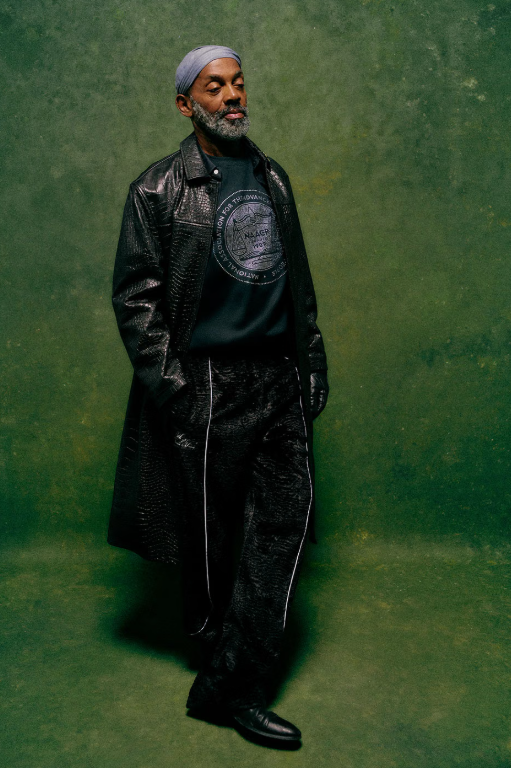 A bearded man wearing a black leather jacket and hat stands in front of a green background.