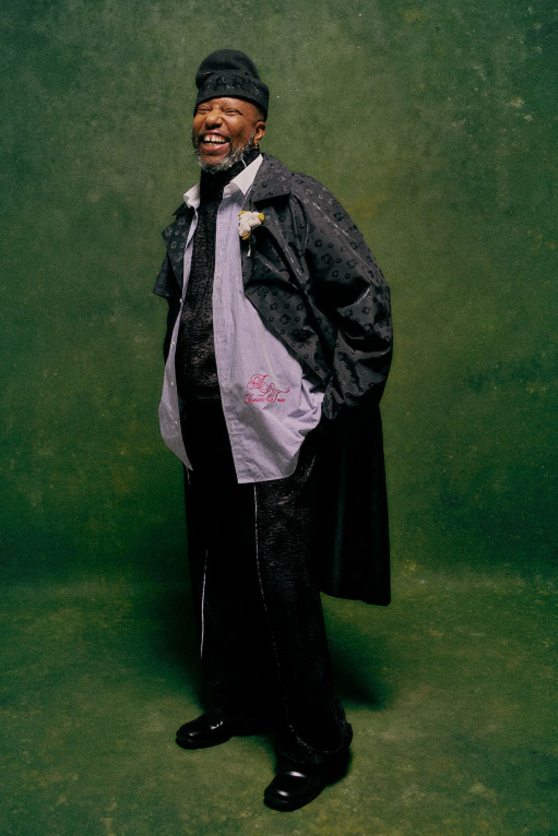 A smiling older man wearing a black hat and coat stands against a green background.