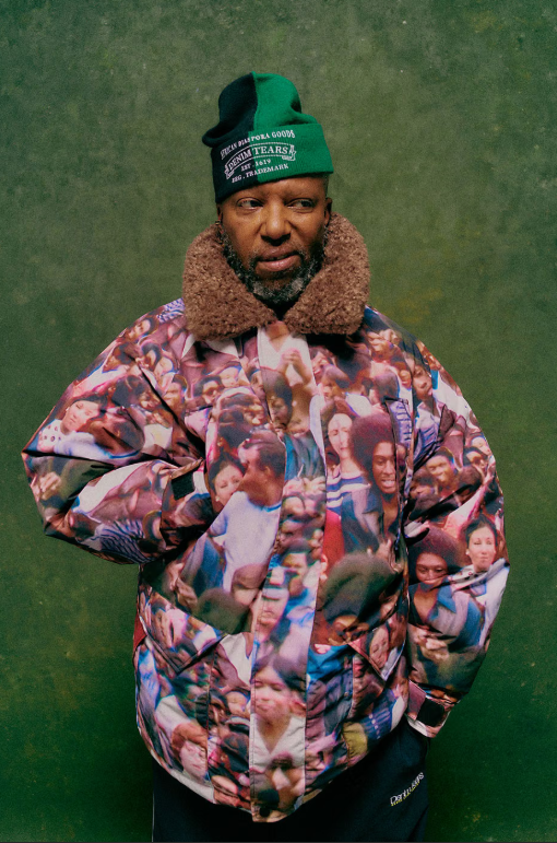 A man wearing a green hat and a furry jacket with a colorful, crowded pattern stands against a green background.