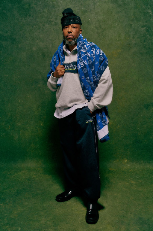 A man wearing a blue and white patterned jacket, gray sweatshirt, and black hat stands in front of a green background.