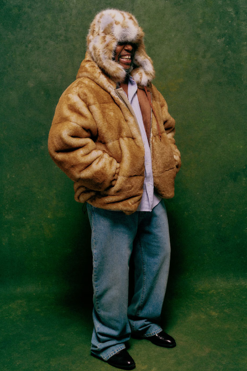 A person wearing a large, furry tan coat with a hood covering their face, standing against a green background.