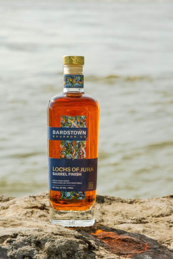 A bottle of Bardstown Bourbon Co. "Lochs of Jura Barrel Finish" whiskey on a rocky beach with a body of water in the background.