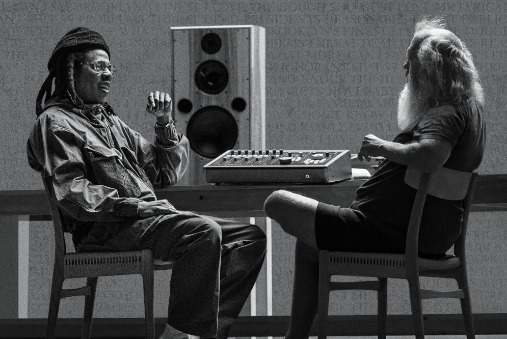 Two men sitting at a sound mixing console, one wearing a hat and glasses, the other with long hair.