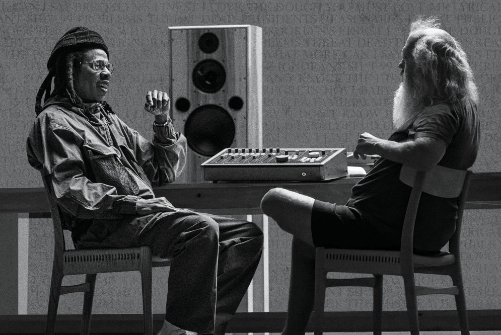 Two men sitting at a sound mixing console, one wearing a hat and glasses, the other with long hair.