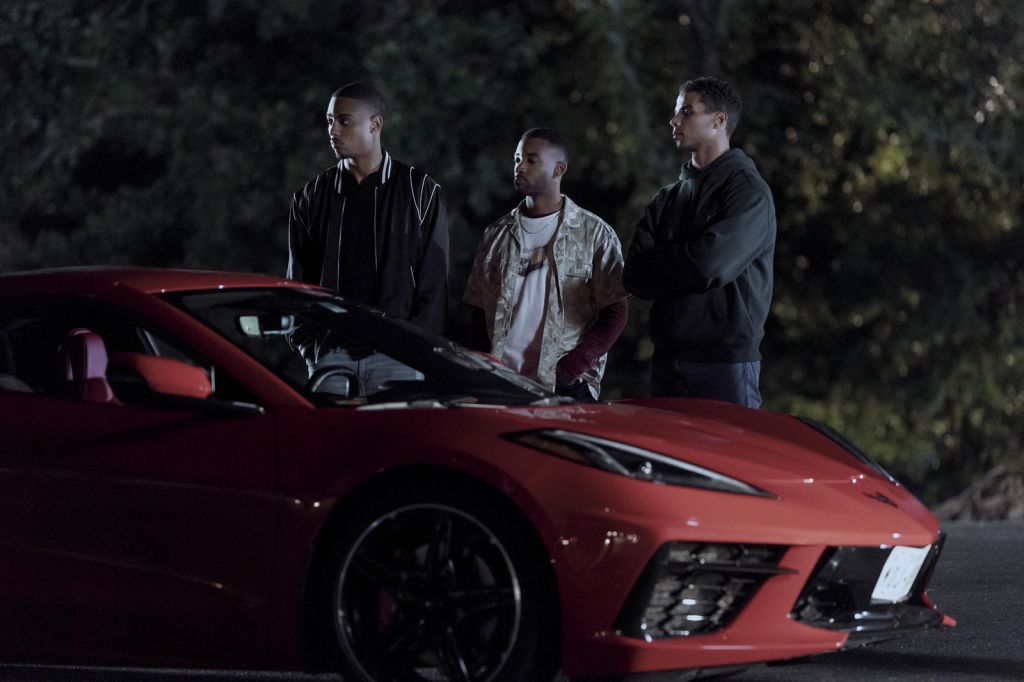 Three young men standing near a red sports car in a dark outdoor setting.